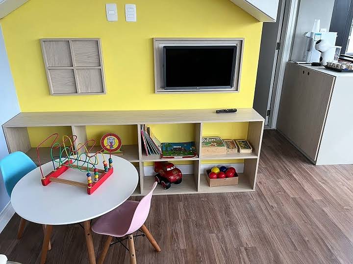 Espaço infantil com parede amarela em formato de casinha, estante de brinquedos e mesinha